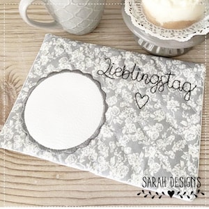 May include: A gray and white fabric coaster with a white circle in the center and the text "Lieblingstag" with a heart above it. The text "SARAH DESIGNS" is at the bottom of the coaster with a small heart and vine design.