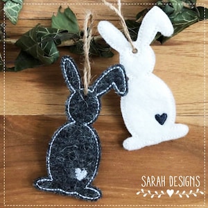 Könnte beinhalten: Zwei Filz-Hasen-Anhänger, einer schwarz und einer weiß, mit einem kleinen weißen Herz auf jedem. Die Hasen hängen an einer Schnur.