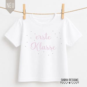 Peut inclure: Un t-shirt blanc avec l'inscription "erste Klasse" en rose, entourée de petits points gris. Le t-shirt est suspendu à une corde à linge avec des pinces à linge en bois. Le texte "SARAH DESIGNS" est en bas.