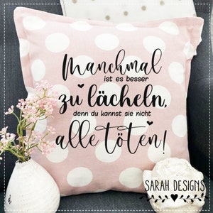 Puede incluir: Cojín rosa con lunares blancos y texto negro que dice "Manchmal ist es besser zu lächeln, denn du kannst sie nicht alle töten!"  El cojín también tiene el texto "SARAH DESIGNS" en negro con un corazón.