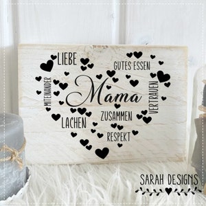 Könnte beinhalten: Ein weißes Holzschild mit einer Herzform aus schwarzen Herzen und dem Wort "Mama" in der Mitte. Das Herz ist umgeben von Wörtern wie "Liebe", "Gutes Essen", "Zusammen", "Respekt", "Lachen", "Vertrauen" und "Miteinander". Am unteren Rand des Schildes steht "SARAH DESIGNS" mit einem kleinen Blumenmuster.