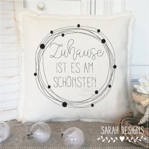 Könnte beinhalten: Ein weißes Kissen mit schwarzem Text, der "Zuhause ist es am schönsten" (Zuhause ist es am schönsten) lautet, und einem schwarzen Kreisdesign mit kleinen schwarzen Punkten drumherum. Das Kissen liegt auf einer weißen Oberfläche mit einem Holzhintergrund.