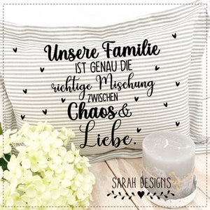 May include: A grey and white striped cushion with black text that reads "Unsere Familie ist genau die richtige Mischung zwischen Chaos & Liebe." The cushion is sitting on a wooden surface with a white flower and a grey candle.