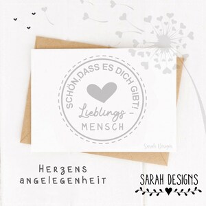 May include: A white greeting card with a grey heart and the words "SCHÖN, DASS ES DICH GIBT! Lieblings MENSCH" in a circular design. The card is inside a brown envelope. The text "HERZENS ANGELEGENHEIT" is below the card.