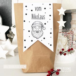 Könnte beinhalten: Ein brauner Papiergeschenktüte mit einem weißen Banner mit einer Weihnachtsmann-Illustration und dem Text "vom Nikolaus". Das Banner ist mit schwarzen Sternen verziert. Weihnachtliches Thema.