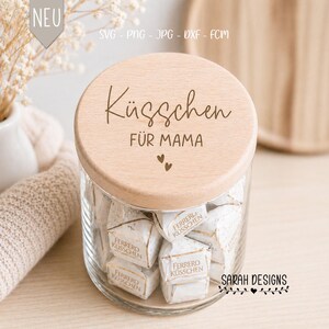 Könnte beinhalten: Ein klares Glas mit einem Holzdeckel, gefüllt mit einzeln verpackten Pralinen. Auf dem Deckel stehen die Worte "Küsschen FÜR MAMA" und zwei kleine Herzen. Das Glas ist mit "SARAH DESIGNS" beschriftet.