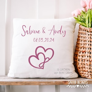 May include: White pillow with pink lettering that says "Sabine & Andy 08.09.2021" and two pink intertwined hearts. The pillow also has the text "13 Größen von 8cm - 20cm" and the text "SARAH DESIGNS" at the bottom.