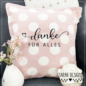 Könnte beinhalten: Ein rosa Kissen mit weißen Punkten und dem Text "danke für alles" in schwarzer, kursiv geschriebener Schrift. Das Kissen ist mit einem kleinen Herzsymbol verziert.