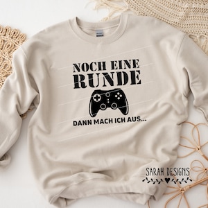 Puede incluir: Sudadera beige con el texto alemán "NOCH EINE RUNDE" sobre un gráfico de mando negro y el texto "DANN MACH ICH AUS..." debajo. La sudadera es de color neutro, adecuada para uso casual.