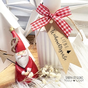 May include: A festive gift tag with the words "LIEBE Weihnachtsgrüße" and a Christmas tree is tied with a red and white gingham ribbon. A small Santa Claus figurine and a white candle are also visible.