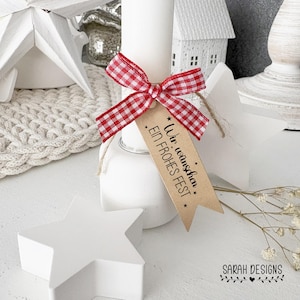 May include: A white candle adorned with a red and white gingham ribbon and a tag reading "Wir wünschen EIN FROHES FEST". The candle is on a white base, with white star decorations. The scene evokes a festive atmosphere, suitable for seasonal decor.