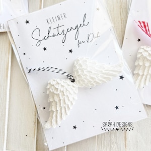 May include: White angel wings attached to a card with the text "Kleiner Schutzengel für Dich" and star graphics. The wings are made of a textured material and are connected to the card with a black and white string. The card is in a clear plastic sleeve.