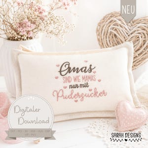 Puede incluir: Un cojín rectangular blanquecino con texto bordado en marrón y rosa. El texto dice "Omas sind wie Mamas nur mit Puderzucker." Una decoración en forma de corazón rosa y un jarrón de flores están en el fondo.