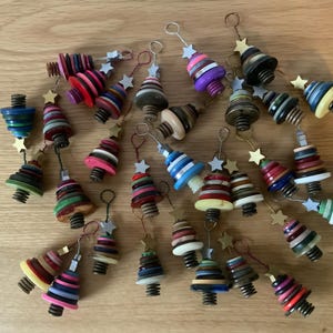 Könnte beinhalten: Eine Sammlung handgefertigter Weihnachtsbaumschmucke. Jeder Baum besteht aus gestapelten, farbenfrohen Knöpfen und ist mit einem kleinen goldenen Stern versehen. Die Ornamente variieren in Farbe und Knopfgröße, mit einer Metallschlaufe zum Aufhängen.
