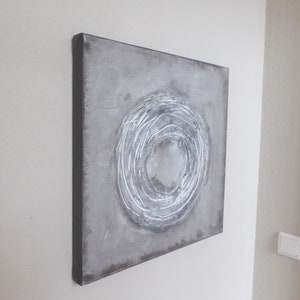 Könnte beinhalten: Quadratische Leinwandkunst mit grauem Hintergrund und einem weißen, strukturierten Design aus konzentrischen Kreisen. Das Kunstwerk ist auf einem schwarzen Rahmen montiert und hängt an einer weißen Wand.