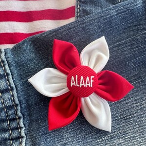 Könnte beinhalten: Eine Stoffblumenbrosche mit roten und weißen Blütenblättern und einem roten Mittelteil. In der Mitte der Blume ist das Wort "ALAAF" in Weiß gestickt. Die Brosche ist auf einer Denim-Oberfläche befestigt.