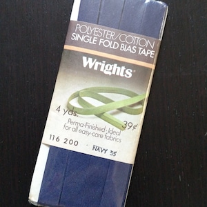 Op de afbeelding: Een verpakking met 4 yards marineblauwe enkelvoudige vouw biaisband van Wrights. De verpakking is gelabeld met "Polyester/Katoen Enkelvoudige Vouw Biaisband" en "Perma-Finished: Ideaal voor alle onderhoudsvriendelijke stoffen".