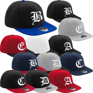 Könnte beinhalten: Eine Sammlung von Snapback-Baseballkappen in verschiedenen Farben, darunter Schwarz, Rot, Marineblau und Grau. Jede Kappe hat einen weißen, gestickten Buchstaben in gotischer Schrift auf der Vorderseite, mit einem kontrastierenden Schirm.