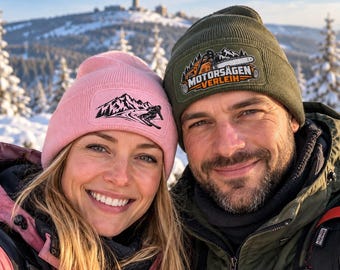 May include: A light pink beanie with a black mountain and skier graphic patch and an olive green beanie with a patch featuring a chainsaw graphic and the text MOTORSÄGEN VERLEIH, both worn in a snowy mountain landscape.