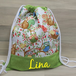 Könnte beinhalten: Ein Kordelzugrucksack mit weißem Hintergrund, der eine farbenfrohe Waldszene mit Eulen, Vögeln und Eichhörnchen zeigt. Der untere und obere Rand sind grün. Der Name "Lina" ist gelb auf dem grünen Boden gedruckt.