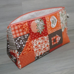 Op de afbeelding: Een patchwork stoffen etui met een verscheidenheid aan oranje, rode, bruine en witte patronen. Het etui is gemaakt van katoenen stof en heeft een ritssluiting.