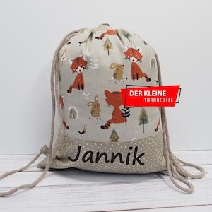 Könnte beinhalten: Turnbeutel mit Kordelzug und hellgrauem Hintergrund mit einem Muster aus roten Pandas, Hasen, Bäumen und Regenbogen. Der untere Teil ist hellgrau mit weißen Punkten und dem Namen "Jannik" in Schwarz. Der Text "DER KLEINE TURNBEUTEL" befindet sich auf einem roten Banner.