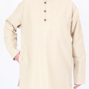 May include: Beige long-sleeve tunic with a mandarin collar and three brown buttons. The tunic has a chest pocket and side slits. The model is wearing white pants. The tunic is made of a light-colored fabric.