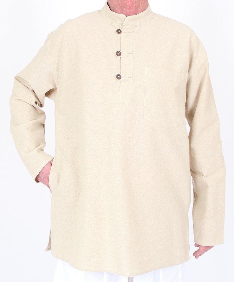 May include: Beige long-sleeve tunic with a mandarin collar and three brown buttons. The tunic has a chest pocket and side slits. The model is wearing white pants. The tunic is made of a light-colored fabric.
