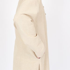 May include: A light beige long-sleeved jacket with a mandarin collar and two buttons. The jacket has two pockets and a side slit. The fabric appears to be a woven material. The jacket is worn over white pants.