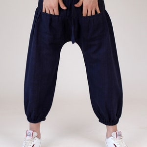 May include: Navy blue harem pants with a drawstring waist and two large front pockets. The pants have a relaxed fit, gathered ankles, and are made of a textured fabric. The person is wearing white sneakers.