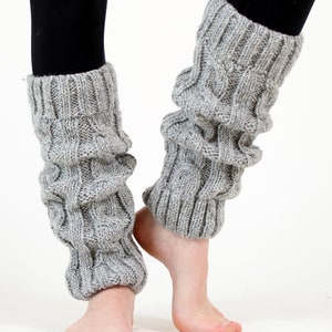 May include: A pair of gray cable knit leg warmers. The leg warmers are made of a soft, textured yarn and have a ribbed cuff at the top and bottom. The leg warmers are shown on a person's legs, with black leggings visible.