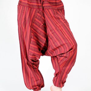 May include: Red and maroon striped harem pants with a drawstring waist and pockets. The loose-fitting pants have a gathered ankle and are made of a lightweight fabric. The vertical stripes create a visually appealing pattern.