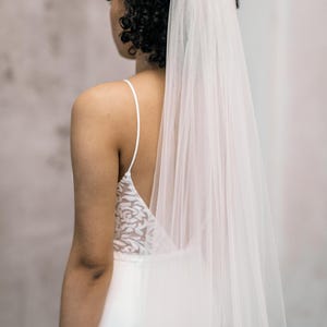 Peut inclure: Une mariée porte une robe de mariée blanche avec un dos nu et des bretelles fines. Un voile blanc transparent tombe dans le dos. La mariée a les cheveux bouclés foncés. La robe a un motif de dentelle florale dans le dos.