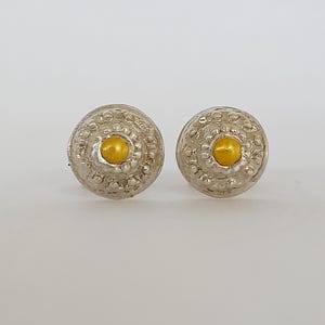 Peut inclure: Une paire de boucles d'oreilles à clous argentées avec un motif texturé. Chaque boucle d'oreille présente un petit centre rond de couleur or. Les boucles d'oreilles sont posées sur un fond clair et uni.