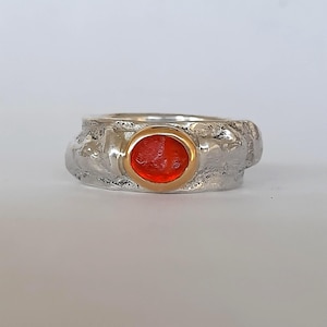 Op de afbeelding: Een zilveren ring met een getextureerde band en een gouden bezel setting. De ring heeft een levendige, ovaalvormige rode edelsteen. Het ontwerp is eenvoudig maar elegant, geschikt voor dagelijks gebruik of speciale gelegenheden.
