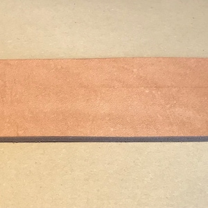 Könnte beinhalten: Ein rechteckiges, hellbraunes Holzstück mit sichtbarer Maserung. Das Holz scheint unbehandelt zu sein und hat eine leicht raue Textur. Die Kanten sind sauber und gerade, was darauf hindeutet, dass es für einen bestimmten Zweck geschnitten wurde. Das Holz ist etwa 2.5 cm dick.