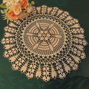 Può includere: Un centrino circolare color crema con un intricato design all'uncinetto. Il centrino presenta un motivo floreale centrale circondato da cerchi concentrici e un bordo smerlato. Un bouquet di fiori color pesca e bianchi poggia in alto a sinistra.