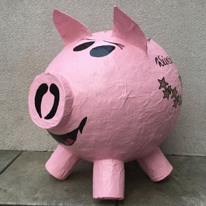 Peut inclure: Une tirelire rose en forme de cochon avec des yeux, un groin et une bouche souriante noirs. La tirelire a quatre pattes et deux oreilles. Le mot "Sweet" est écrit sur le côté, avec un groupe d'étoiles.