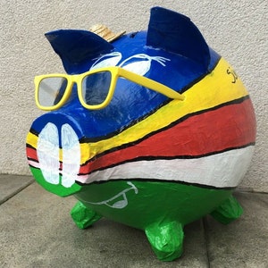 Könnte beinhalten: Ein blaues, gelbes, rotes, weißes und grünes gestreiftes Sparschwein mit einem Strohhut und einer gelben Sonnenbrille. Das Sparschwein hat eine weiße bemalte Schnauze und ein weißes bemaltes Auge.
