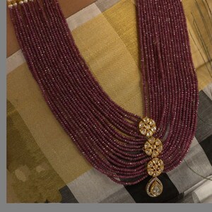 May include: A long, multi-strand necklace made of burgundy beads with gold accents. The necklace features a large, ornate pendant with a teardrop-shaped diamond.