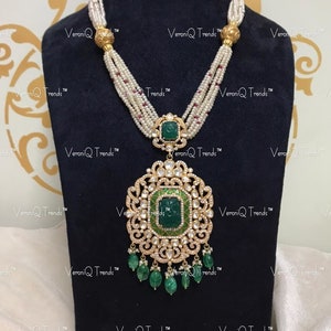 Könnte beinhalten: Eine mehrreihige Perlenkette mit einem großen, verzierten Anhänger. Der Anhänger ist aus Gold gefertigt und hat einen grünen Edelstein, der von kleineren Diamanten umgeben ist. Der Anhänger ist außerdem mit filigranen Verzierungen verziert.