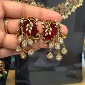 Könnte beinhalten: Ein Paar goldene Ohrringe mit roten und weißen Steinen. Die Ohrringe haben ein florales Design und hängen am Ohr.