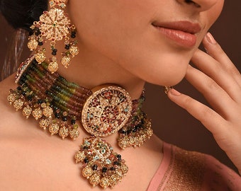 May include: A gold choker necklace with a large round pendant featuring intricate floral designs and colorful gemstones. The necklace is made of multiple strands of beads in shades of green, brown, and black. The pendant is adorned with pearls and red, green, and blue stones.