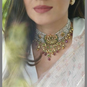 May include: A pearl necklace with a gold pendant, green and red gemstones. The necklace has a matching earring. The jewellery is worn with a white top and a floral patterned fabric.