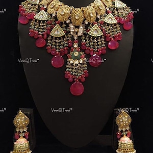 May include: A gold necklace and earrings set with pink and green stones. The necklace features a large pendant with a floral design and is adorned with multiple strands of beads. The earrings are similarly designed with a smaller pendant and multiple strands of beads.