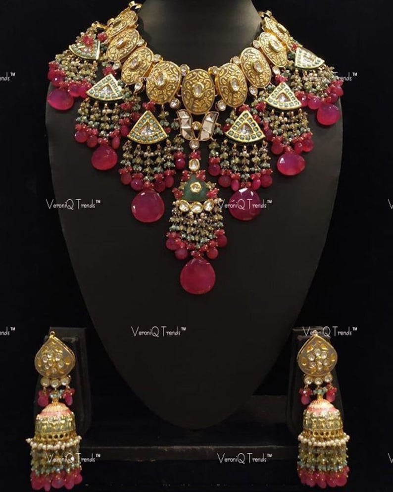 May include: A gold necklace and earrings set with pink and green stones. The necklace features a large pendant with a floral design and is adorned with multiple strands of beads. The earrings are similarly designed with a smaller pendant and multiple strands of beads.