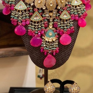 May include: A gold necklace with pink and green stones and a matching pair of earrings. The necklace has a large, ornate pendant with a silver and gold design. The earrings are also ornate and have a similar design to the pendant.