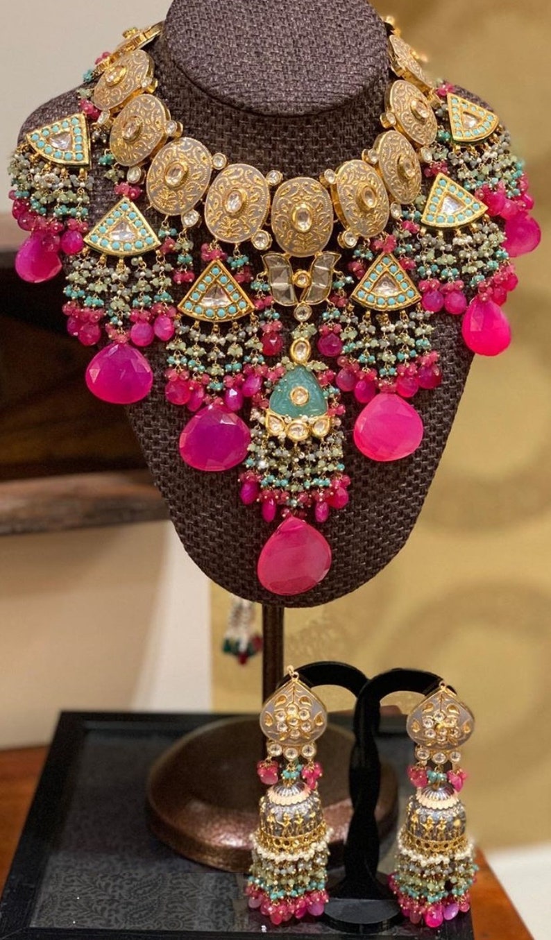 May include: A gold necklace with pink and green stones and a matching pair of earrings. The necklace has a large, ornate pendant with a silver and gold design. The earrings are also ornate and have a similar design to the pendant.