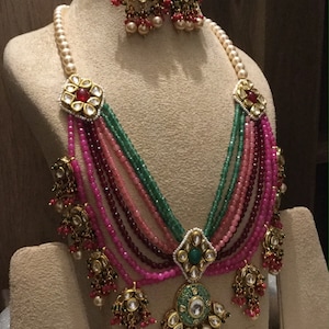 May include: A multi-strand necklace with pink, green, and red beads. The necklace has a large, ornate pendant with a green stone and gold accents. The necklace is displayed on a mannequin with matching earrings.