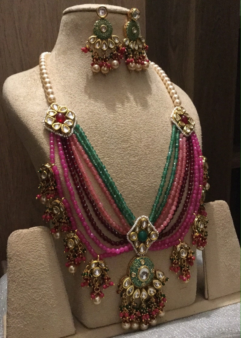 May include: A multi-strand necklace with pink, green, and red beads. The necklace has a large, ornate pendant with a green stone and gold accents. The necklace is displayed on a mannequin with matching earrings.